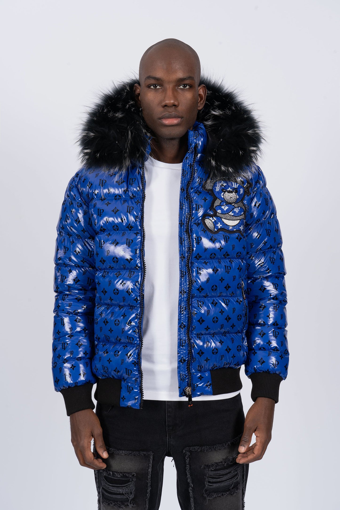 Vista de frente de un modelo con chaqueta plumífero de color azul brillante original con patrón de monograma negro, capucha de pelo oscuro y un gran diseño de un osito gánster con los brazos cruzados en la espalda