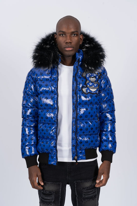 Vista de frente de un modelo con chaqueta plumífero de color azul brillante original con patrón de monograma negro, capucha de pelo oscuro y un gran diseño de un osito gánster con los brazos cruzados en la espalda
