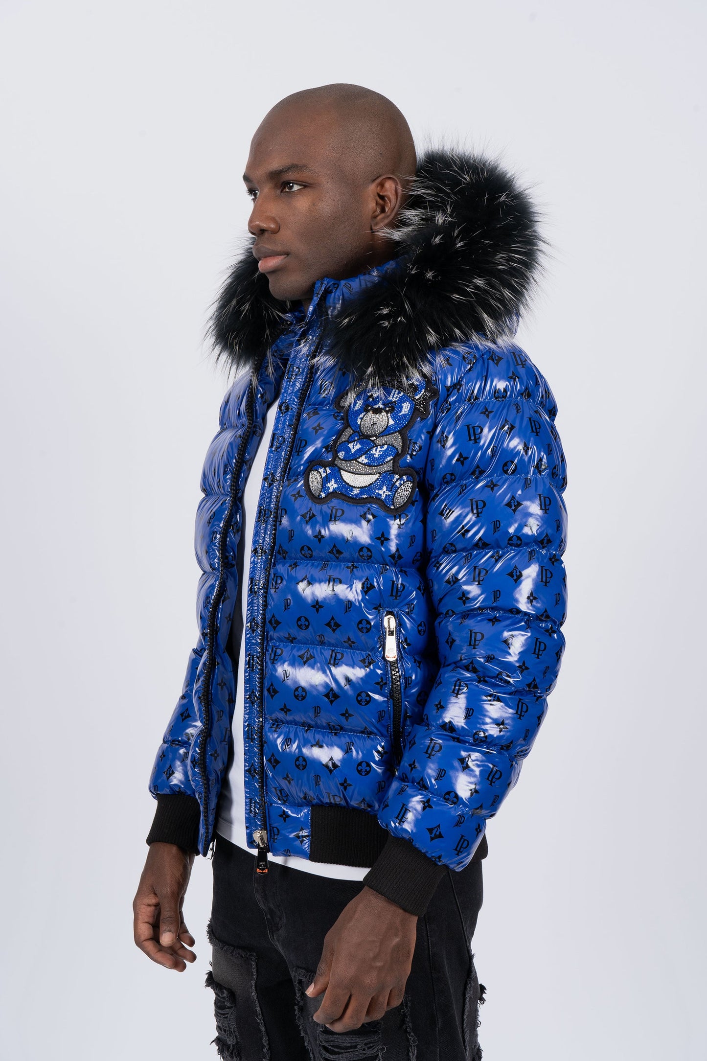 Vista de lado de un modelo con chaqueta plumífero de color azul brillante original con patrón de monograma negro, capucha de pelo oscuro y un gran diseño de un osito gánster con los brazos cruzados en la espalda