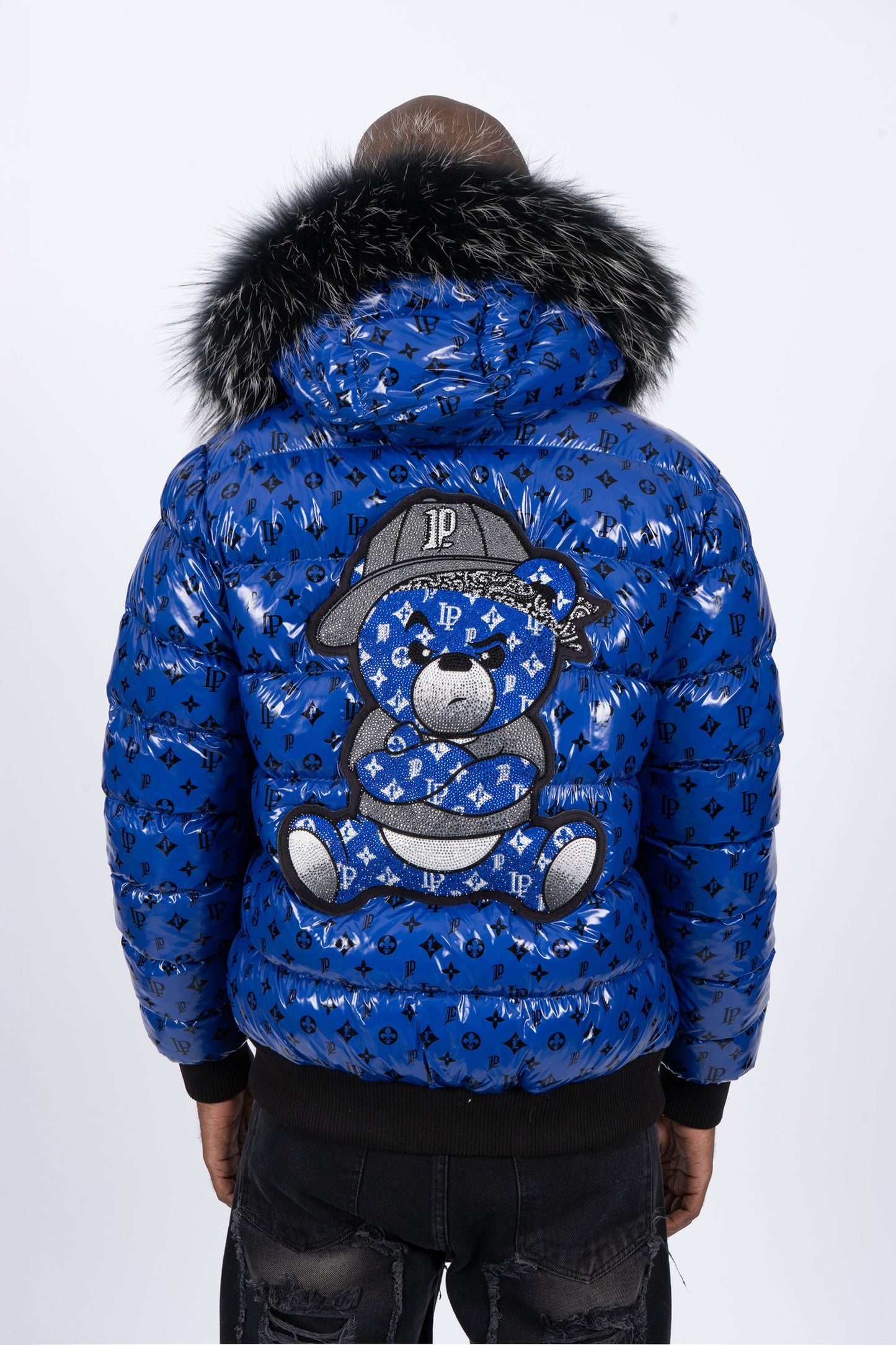 Vista de espaldas de un modelo con chaqueta plumífero de color azul brillante original con patrón de monograma negro, capucha de pelo oscuro y un gran diseño de un osito gánster con los brazos cruzados en la espalda