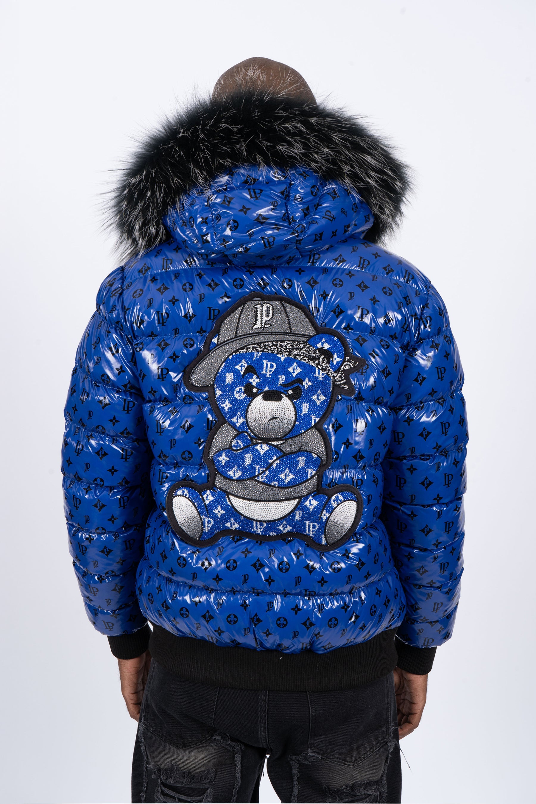Vista de espaldas de un modelo con chaqueta plumífero de color azul brillante original con patrón de monograma negro, capucha de pelo oscuro y un gran diseño de un osito gánster con los brazos cruzados en la espalda