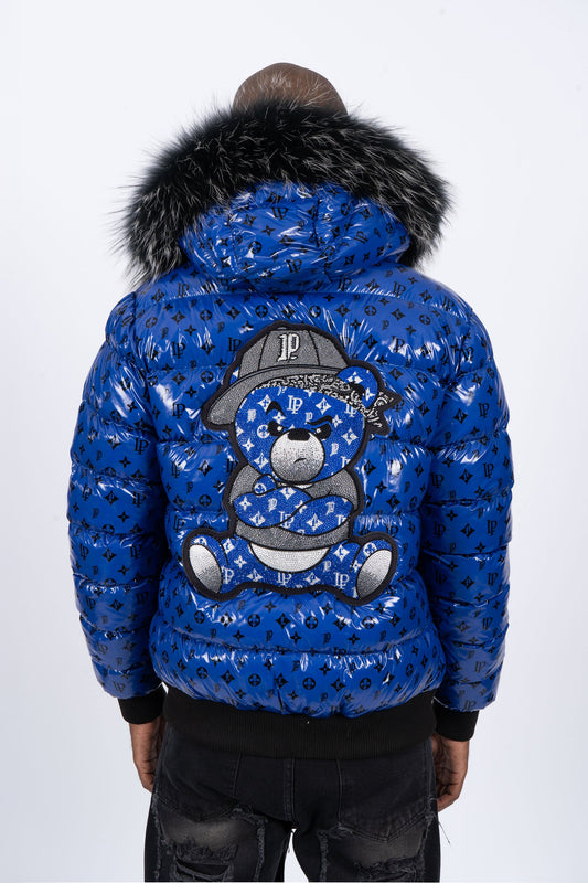 Vista de espaldas de un modelo con chaqueta plumífero de color azul brillante original con patrón de monograma negro, capucha de pelo oscuro y un gran diseño de un osito gánster con los brazos cruzados en la espalda